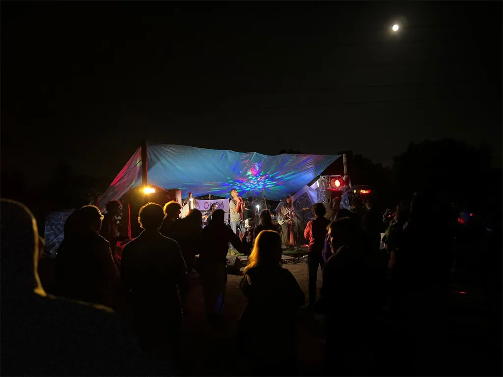 A crowd at night in front of a small lit stage under a canopy